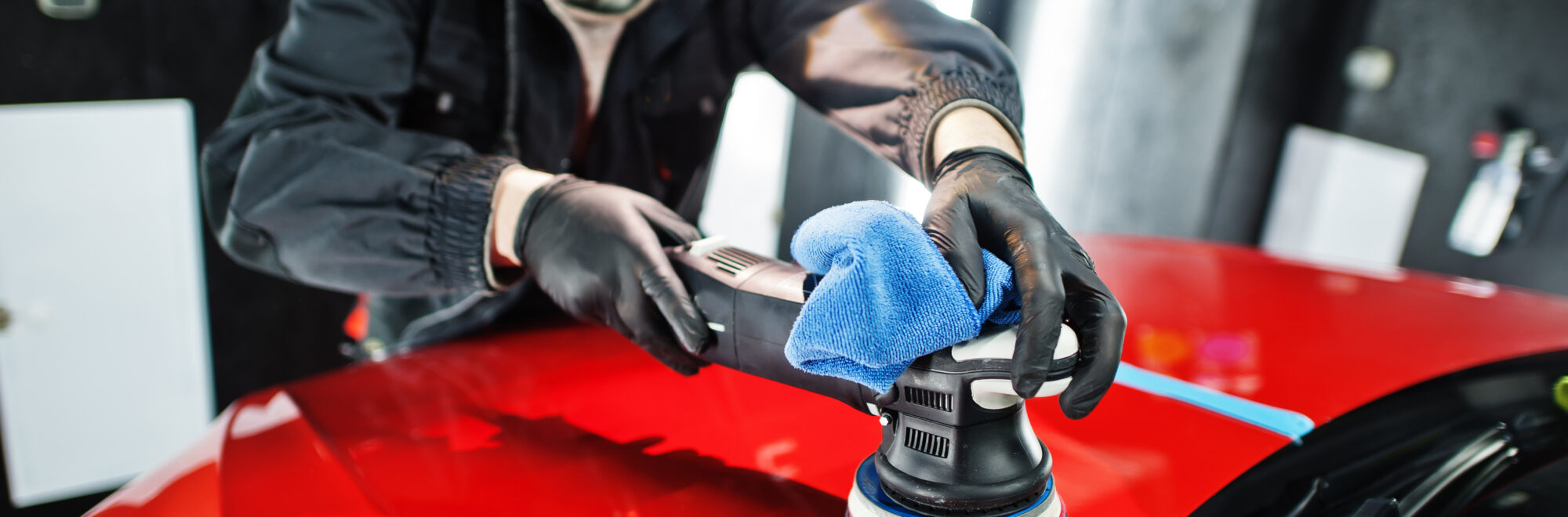 Car detailing concept. Man in face mask with orbital polisher in repair shop polishing orange suv car.